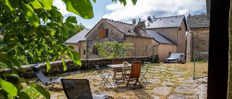 Une terrasse en pierre avec des meubles d’extérieur, un bâtiment en pierre avec un toit en tuiles et une voiture garée à proximité.