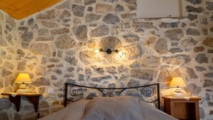 Slaapkamer met een stenen muur, een bed met een gebreid deken, twee nachtkastjes met lampen en een houten plafond.