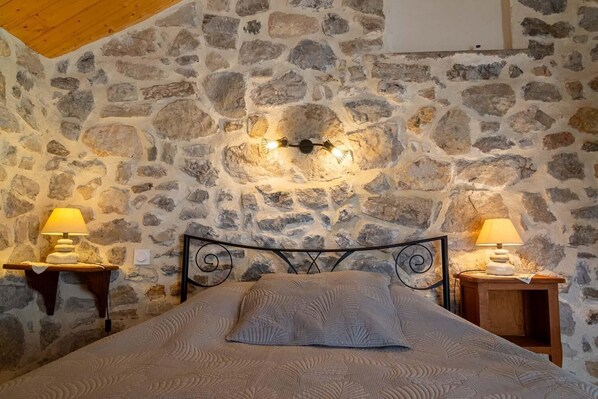 A bedroom with a stone wall, a bed with a textured blanket, two bedside tables with lamps, and a wooden ceiling.
