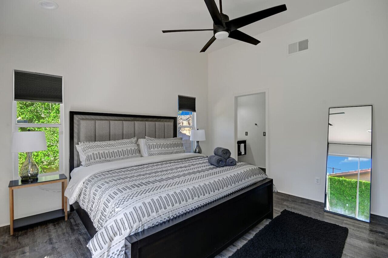 Une chambre moderne avec un grand lit, un ventilateur de plafond, une fenêtre avec des stores, une table de chevet avec une lampe et une vue sur la verdure à l’extérieur.
