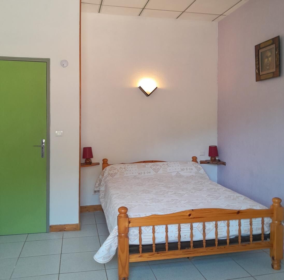 Une chambre avec un lit en bois, une porte verte, une lampe fixée au mur et une petite étagère sur laquelle se trouve une lampe.