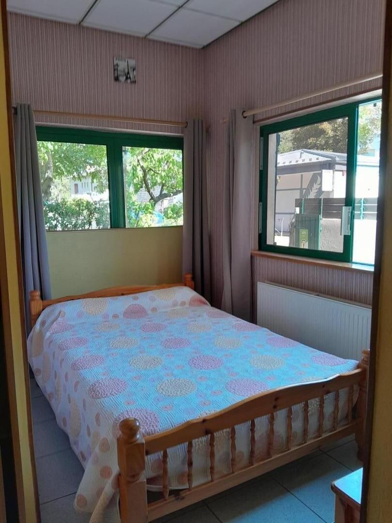 Un lit simple avec une couvre-lit à motifs, dans une pièce dotée de deux fenêtres, dont l’une offre une vue sur les arbres et les bâtiments à l’extérieur.