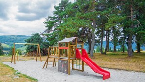 A playground with a wooden slide and swings, set on sandy ground with trees and hills in the background.