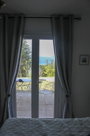 A room with a bed, a window with curtains, a view of a garden and mountains.