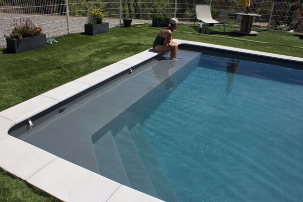 A swimming pool with a person sitting on the edge, surrounded by a well-maintained lawn and a fence.