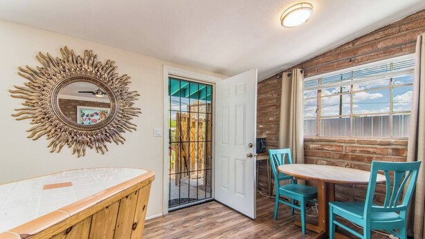 Una habitación con un banco de madera, una mesa redonda con sillas, un espejo con un marco en forma de sol y vistas a un espacio exterior a través de una puerta.