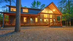 A two-story log cabin with a covered porch, large windows, and a gravel driveway. - Sweet Caroline Creek-Side Cabin 🎯  Games 🔥 Hot Tub 📺 Outdoor TV (BROKEN BOW)