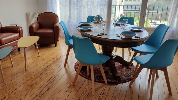 A dining area with a round table, blue chairs, a brown leather sofa, and a small stool. - Grand Logement Rénové, 84 m2, 5 Personnes, à 100 m de la Gare et du Tramway (Le Havre)