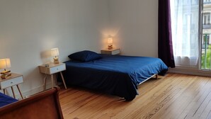 A bedroom with two beds, wooden flooring, bedside tables with lamps, and a window with a view of buildings. - Grand Logement Rénové, 84 m2, 5 Personnes, à 100 m de la Gare et du Tramway (Le Havre)