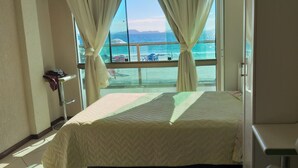 Quarto de hotel com cama, uma cadeira e vista para a praia e oceano através de grandes janelas.
