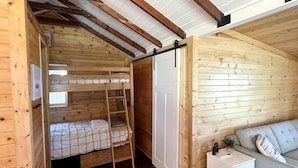 Ein gemütliches Blockhäuschen im Inneren mit Holzausstattung, einem Stockbett, einem kleinen weißen Nachttisch und einer Couch.