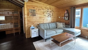 Ein gemütliches Blockhäuschen im Inneren, mit einem Etagenbett, einem grauen Sofa, einem Holztisch und einem Bücherregal.