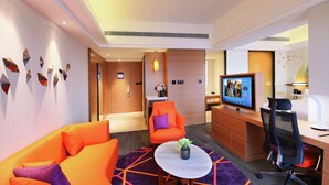 Una habitación de hotel moderna con un sofá, una pequeña mesa redonda con un jarrón, un escritorio con una computadora y un televisor mostrando un programa.