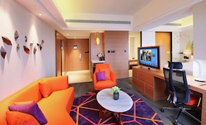 Ein modernes Hotelzimmer mit einer Couch, einem kleinen runden Tisch mit einer Vase, einem Schreibtisch mit einem Computer und einem Fernseher, der ein Programm zeigt.