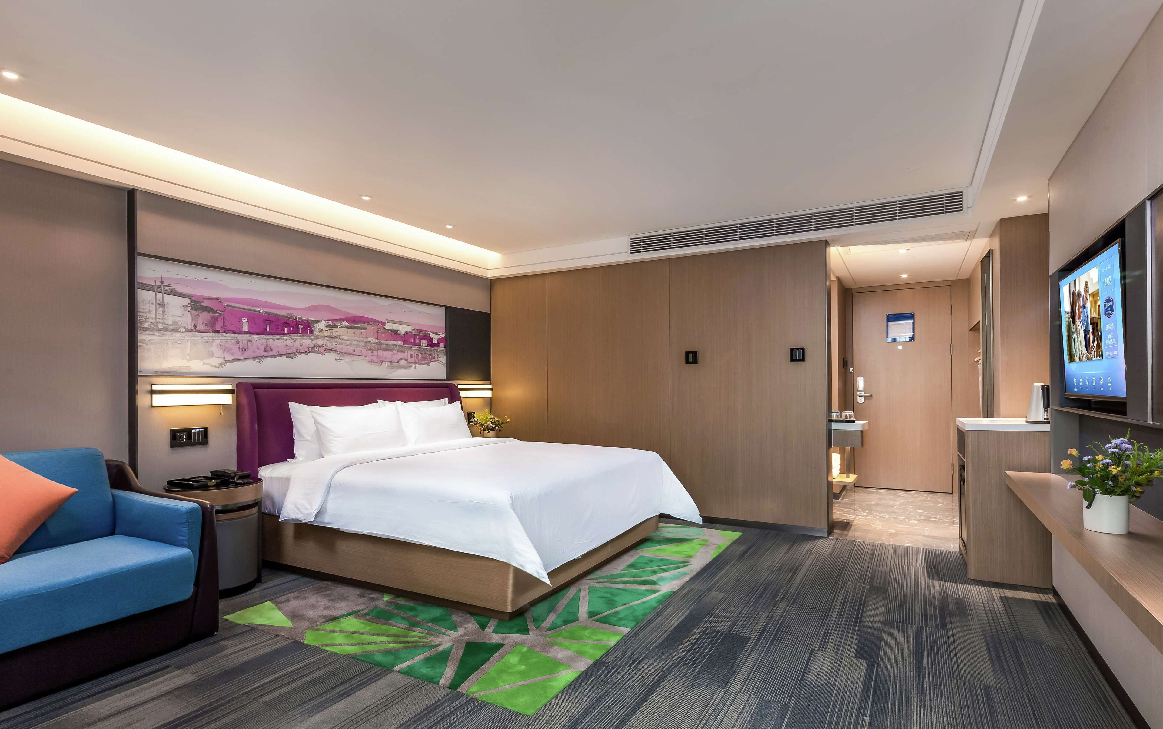 A hotel room with a bed, a nightstand, a blue armchair, a large painting, a TV, and a bathroom visible through an open door.