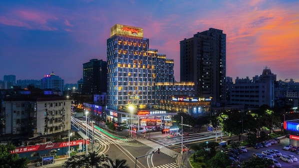 Un hotel de varios pisos con un letrero llamativo en la parte superior, rodeado de otros edificios y un cruce concurrido con tráfico visible.