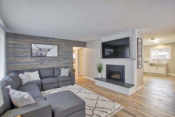 A modern living room with a gray sectional sofa, a fireplace, and a kitchen area in the background.
