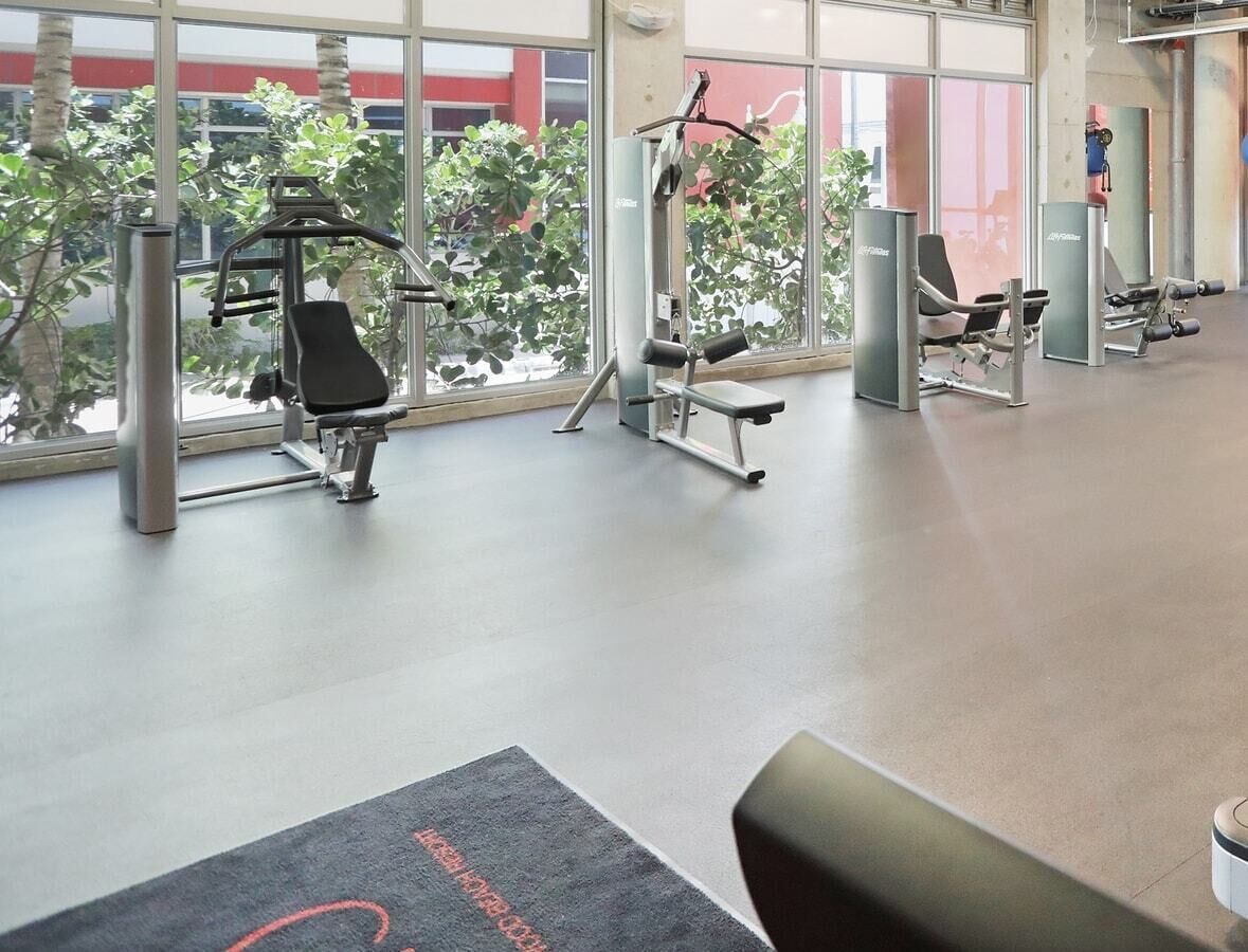 Et fitnesscenter med træningsudstyr, store vinduer og udsigt til træer og bygninger.