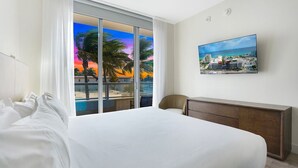 Habitación de hotel con una cama grande, un escritorio y vistas a palmeras y al atardecer a través de ventanales de pared a pared.