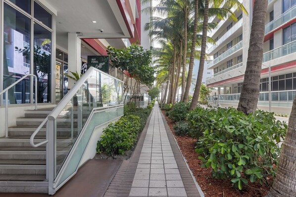 A corridor with a glass railing, palm trees, and a pathway lined with greenery. - Beach Escape Studio I Rooftop Pool (Hollywood)