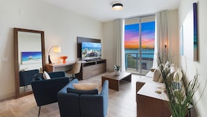 A modern living room with a large TV, a sofa, a desk, and a view of the sunset over the water. - Luxury Waterfront Condo w/ Stunning Sunset View-CS36 (Hollywood)