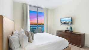 A hotel room with a bed, a TV, a wooden dresser, and a view of the sunset over the water. - Luxury Waterfront Condo w/ Stunning Sunset View-CS36 (Hollywood)
