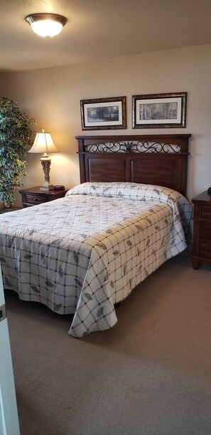 A bedroom with a bed, a nightstand with a lamp, two framed pictures on the wall, and a potted plant.