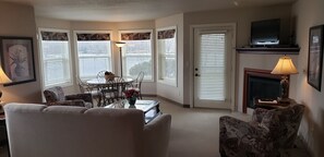 A living room with a sofa, patterned chairs, a dining table with chairs, a fireplace, and a television on a stand. - Unit 316 1 Bdrm/ 1 Bath Condo (Birchwood)
