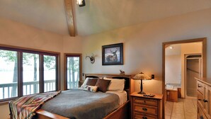 Une chambre à coucher avec un grand lit, une table de chevet, une lampe, un tableau représentant un cerf encadré et une fenêtre donnant sur des arbres.