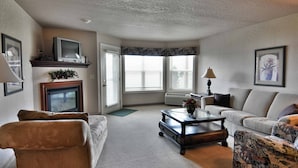 A living room with a fireplace, a sofa, a patterned armchair, and a television on a stand. - Unit 312 1 BD/ 1 BA Condo (Birchwood)