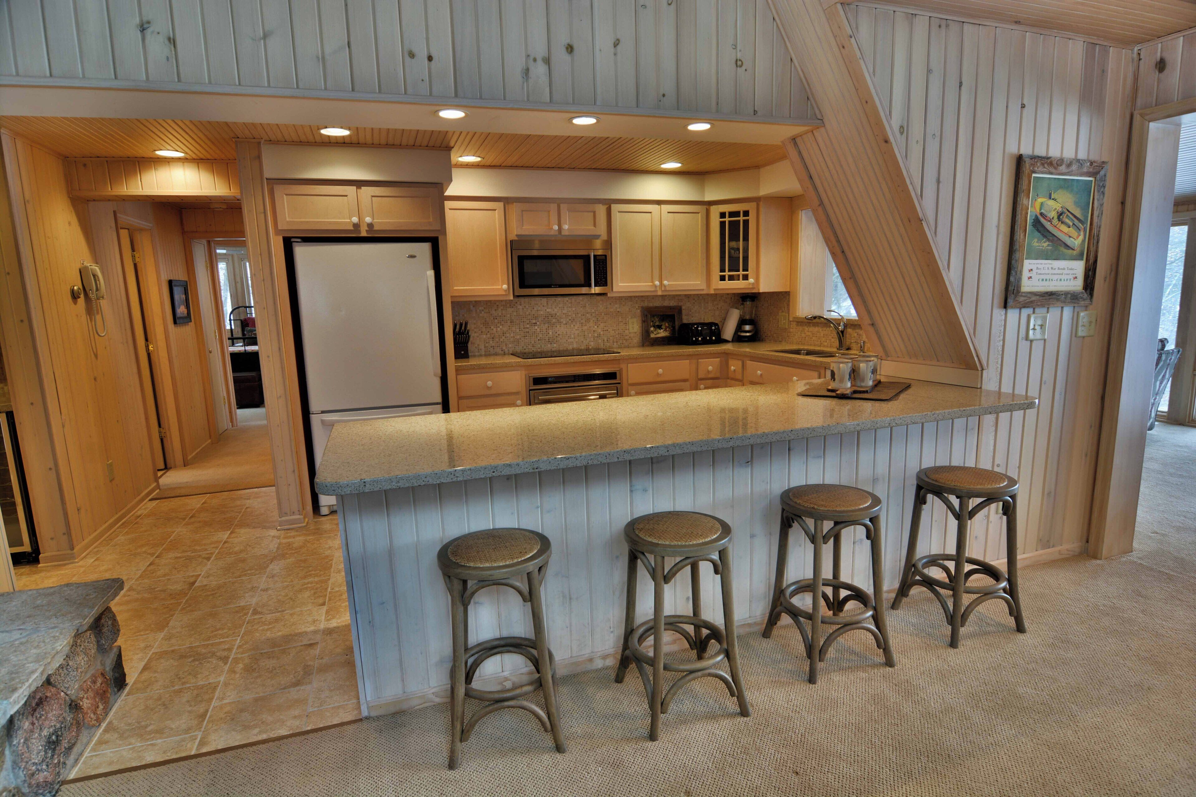 A kitchen with wooden cabinets, a microwave, a refrigerator, and a bar area with stools.