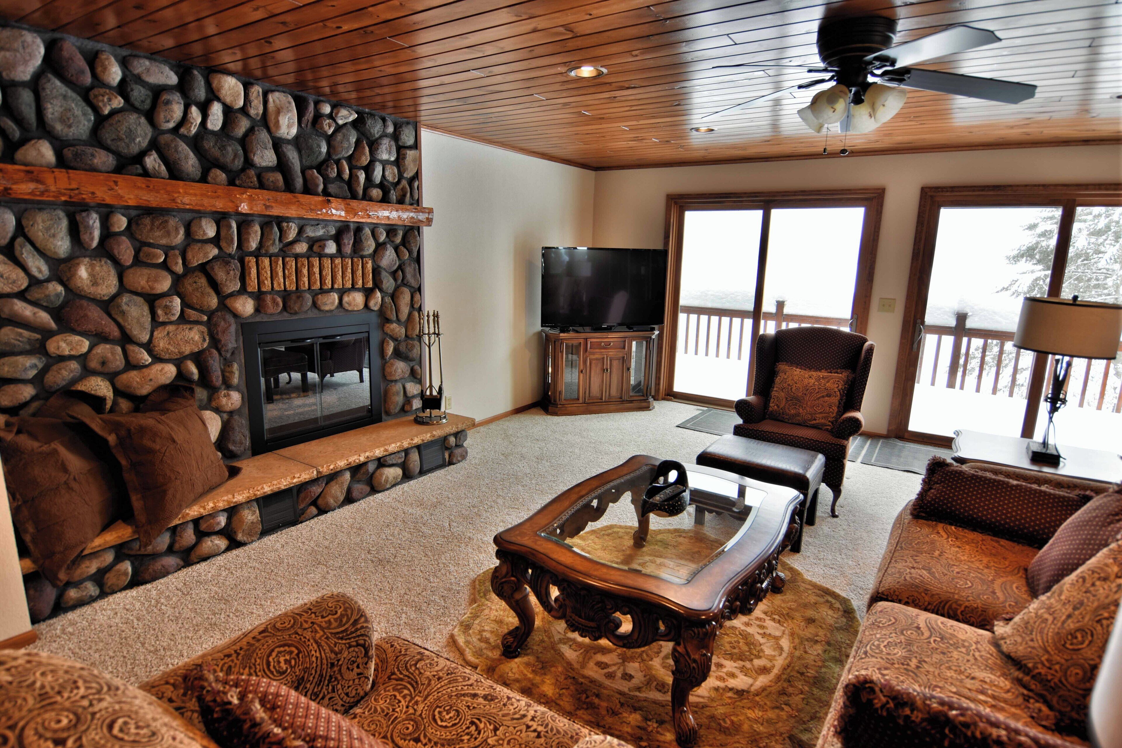 A cozy living room with a stone fireplace, comfortable seating, a flat-screen TV, and a ceiling fan.