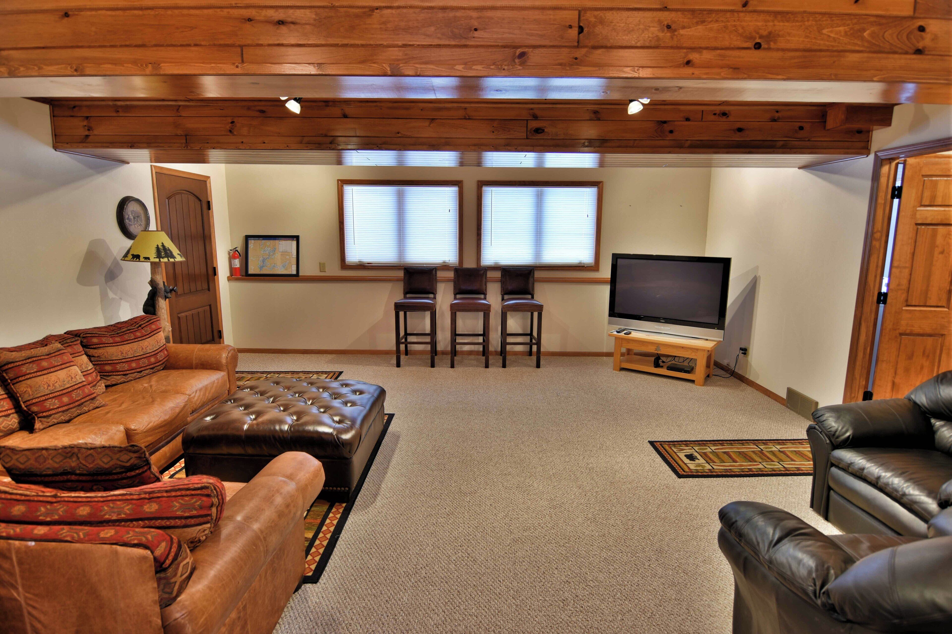 A cozy living room with a leather sofa, a matching ottoman, and a television on a wooden stand. There are two windows with blinds, a door, and a small rug on the floor.