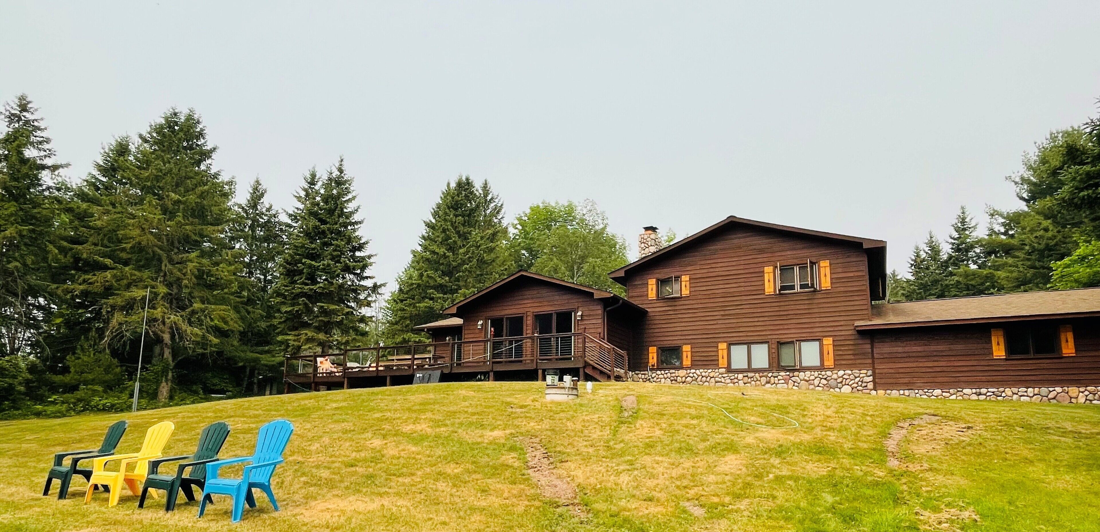 A two-story wooden cabin with a stone foundation, a deck, and a grassy yard with Adirondack chairs.
