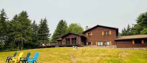 A two-story wooden cabin with a stone foundation, a deck, and a grassy yard with Adirondack chairs.