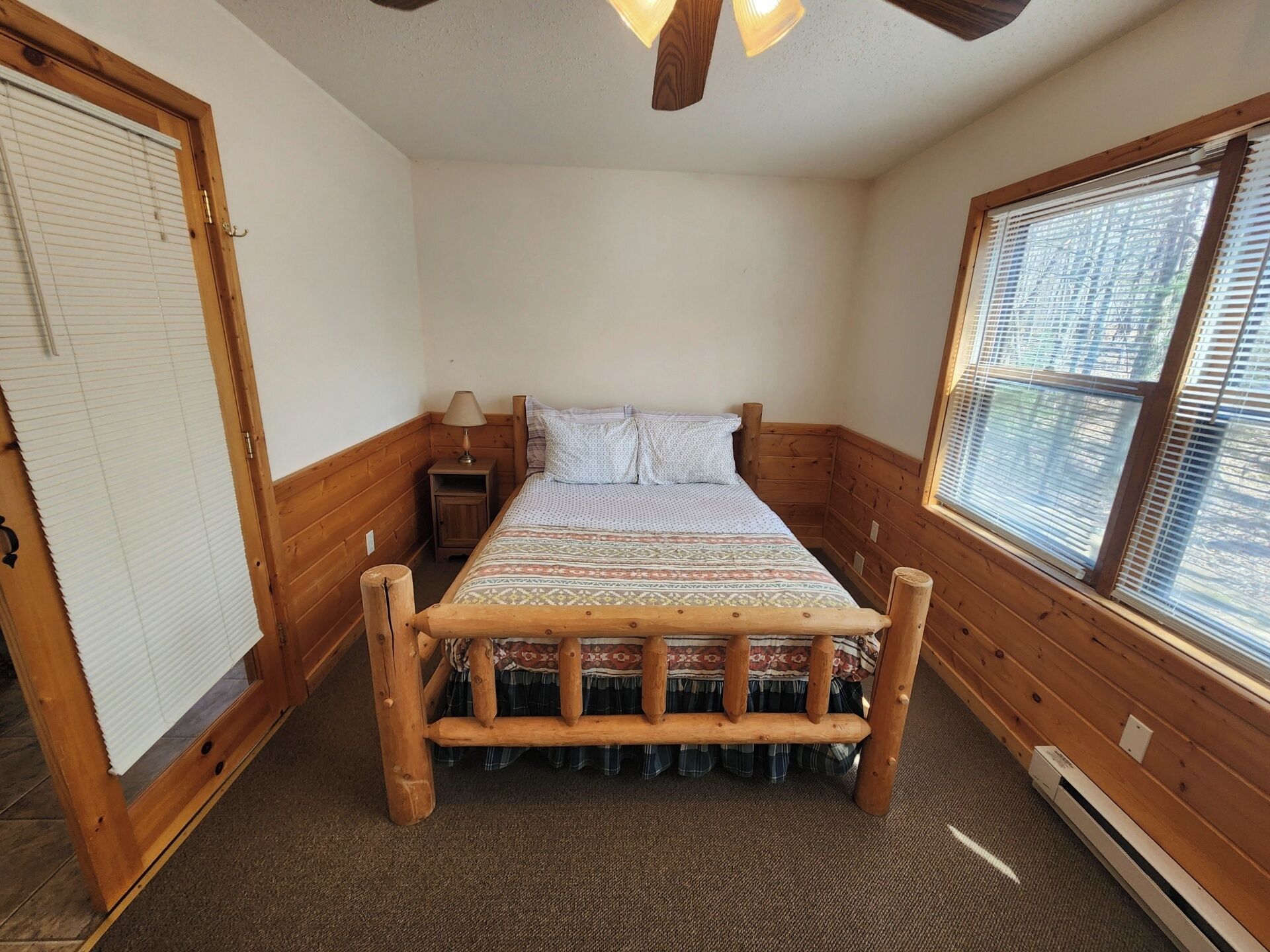 A wooden-framed bed with a patterned bedspread, a nightstand with a lamp, a window with blinds, and a door with a window shade.