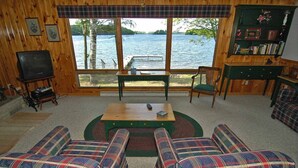 A cozy cabin interior with a large window overlooking a lake, plaid furniture, and a bookshelf. - Seidls (Cable)