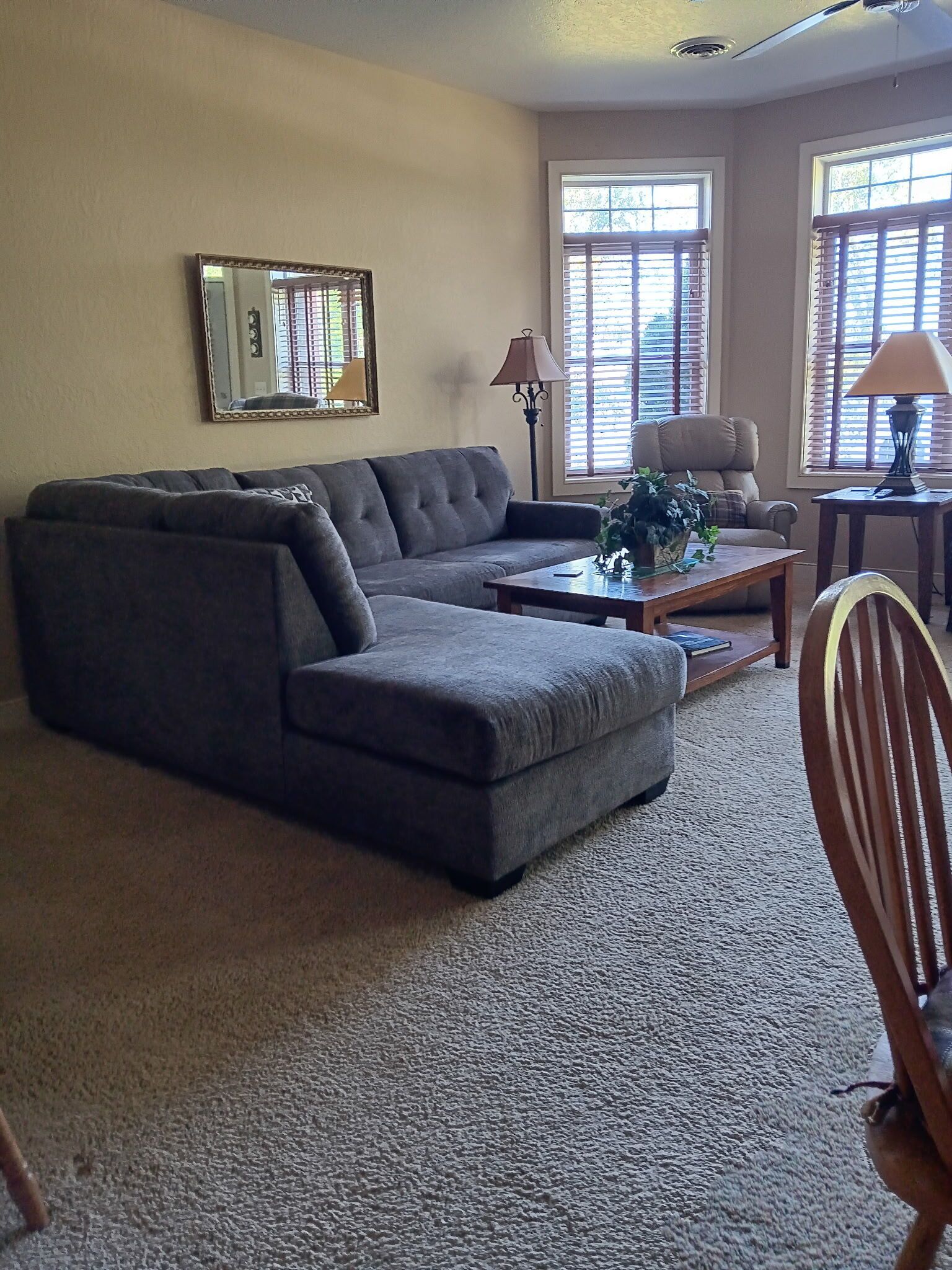 A living room with a sectional sofa, a coffee table, and a chair. There are two windows with blinds, a lamp, and a mirror on the wall.