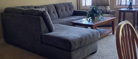 A living room with a sectional sofa, a coffee table, and a chair. There are two windows with blinds, a lamp, and a mirror on the wall.