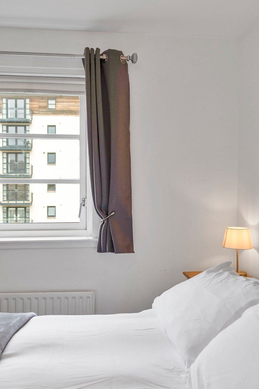 A bedroom with a bed, a window with curtains, a lamp on a bedside table, and a radiator.