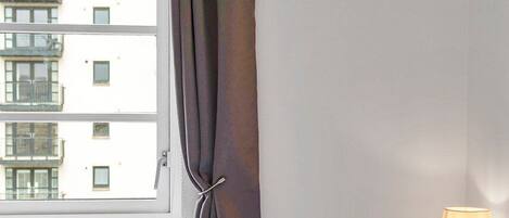 A bedroom with a bed, a window with curtains, a lamp on a bedside table, and a radiator.