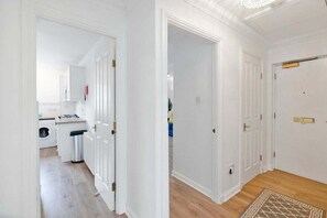 Un couloir aux murs blancs, doté d’un plancher en bois et orné d’un tapis à motifs. Des portes donnent accès à différentes pièces, et on aperçoit à l’arrière-plan un espace dédié à la buanderie.