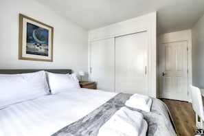 A neatly made bed with white linens and a gray throw blanket, a bedside table with a lamp, a wardrobe, and a door.