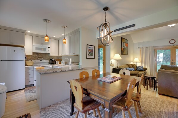 Una cocina moderna con armarios blancos, una mesa de comedor de madera e iluminación colgante.