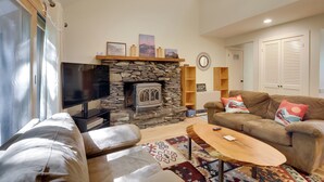 A living room with a stone fireplace, a television, a sofa, a coffee table, and a patterned rug. - Waitsfield Condo w/ Pool & On-Site Trail Access! (Waitsfield)