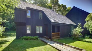 A two-story house with a dark exterior, a front porch, and a wooden ramp leading to the entrance. - Waitsfield Condo w/ Pool & On-Site Trail Access! (Waitsfield)