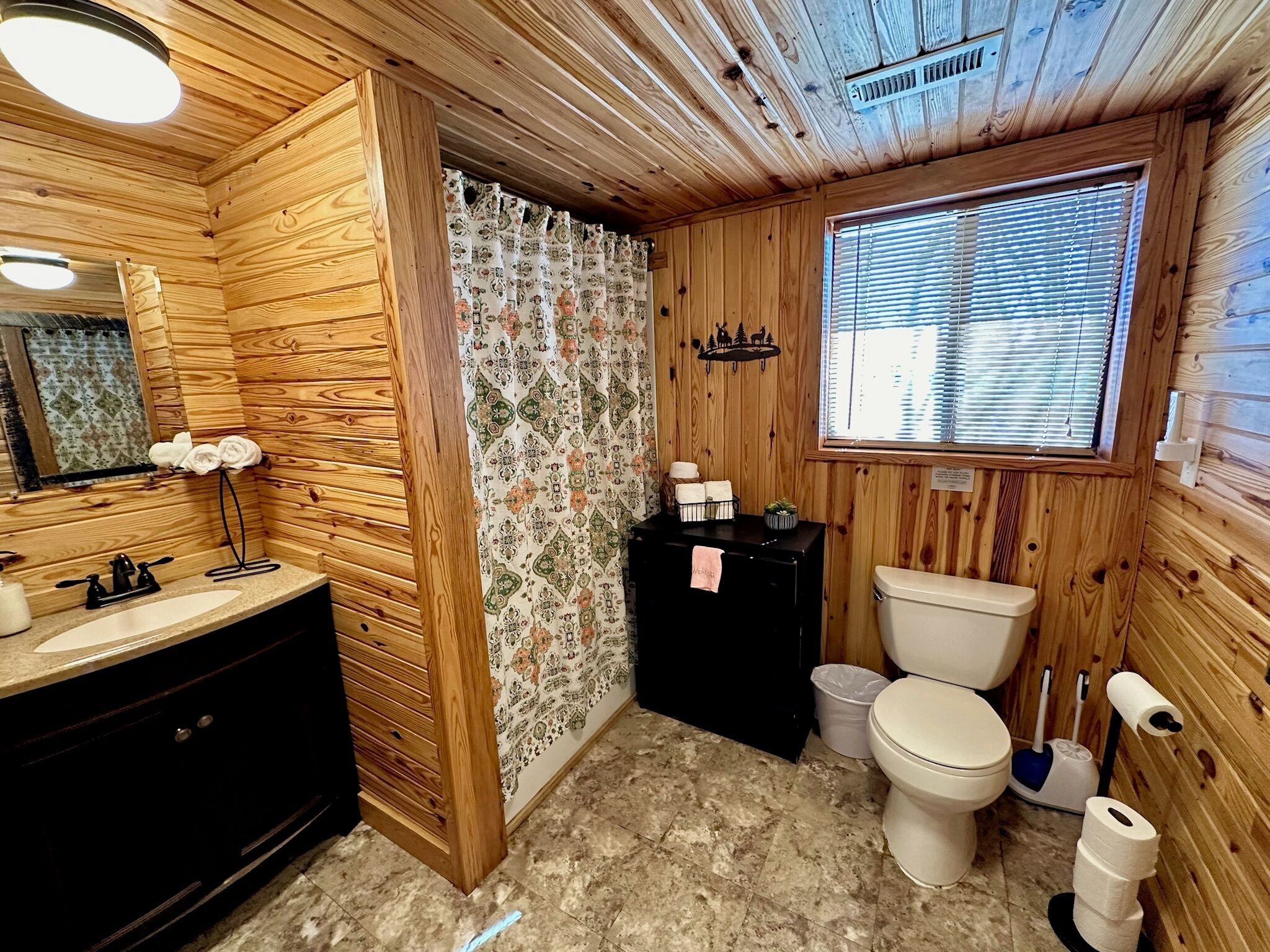 Une salle de bain rustique, avec des murs en bois, un meuble-lavabo en bois, des toilettes et une fenêtre équipée de stores.