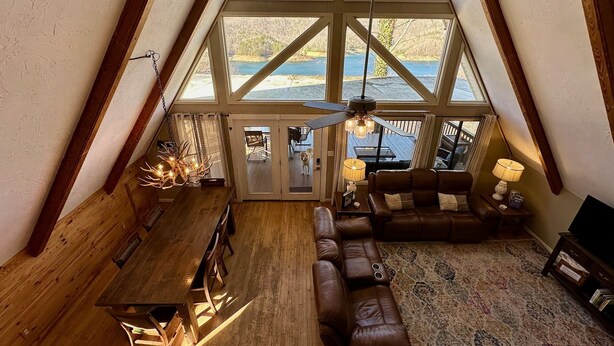 A spacious loft with a high ceiling, large windows offering a view of the outdoors, a wooden dining table, a leather sofa, and a ceiling fan.
