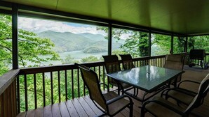 A covered outdoor deck with a table and chairs, overlooking a mountainous landscape and a body of water.