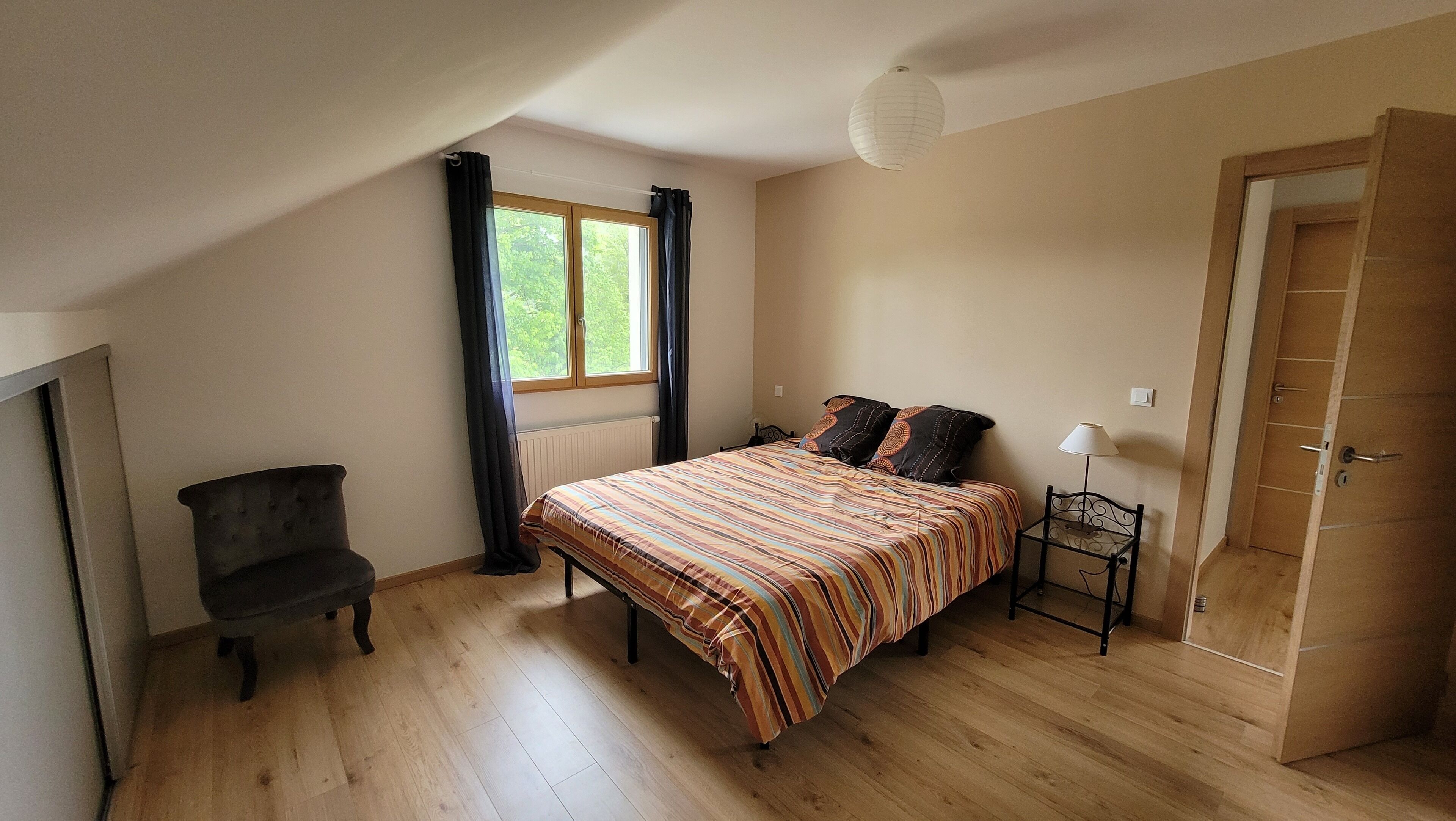 A bedroom with a bed, a chair, a window with curtains, a lamp, and a door.